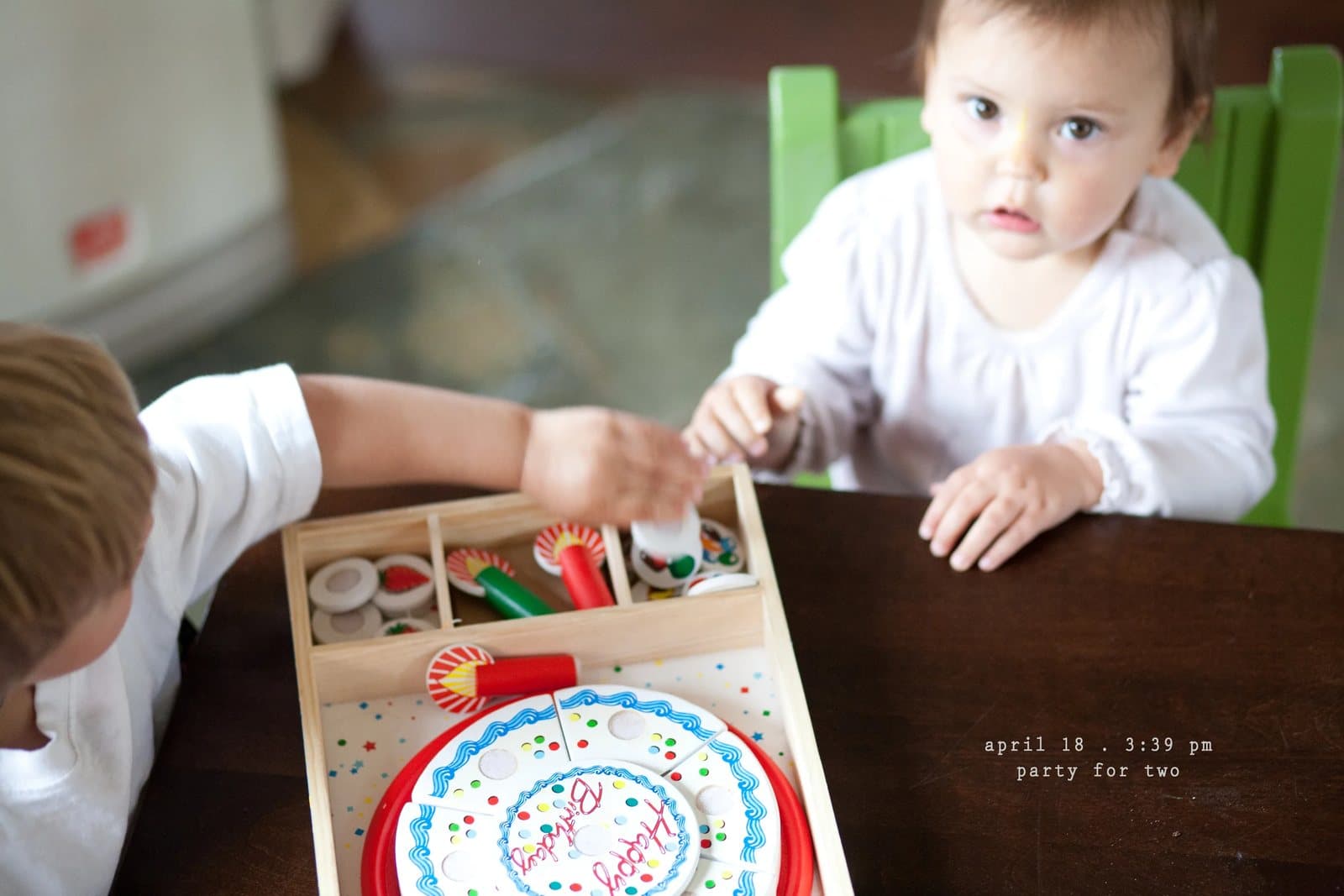 A baby and a boy share a toy birthday cake set at a table, captioned party for two.