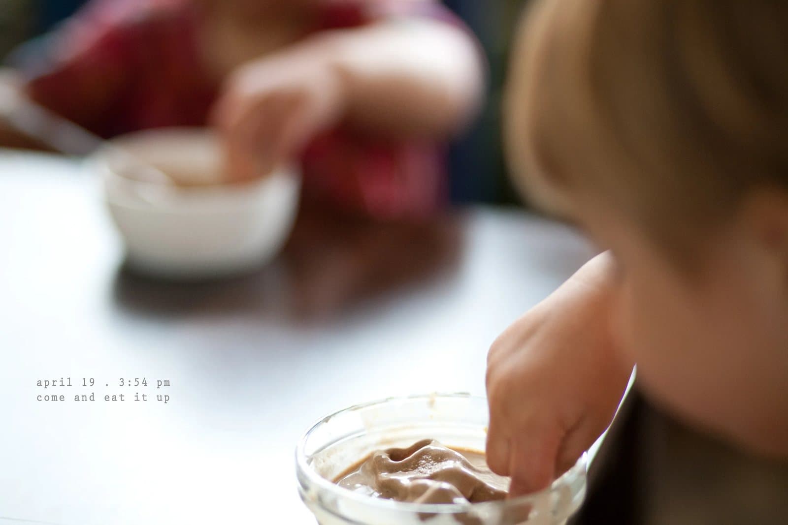 A child dips a finger into a bowl of chocolate pudding, captioned come and eat it up.