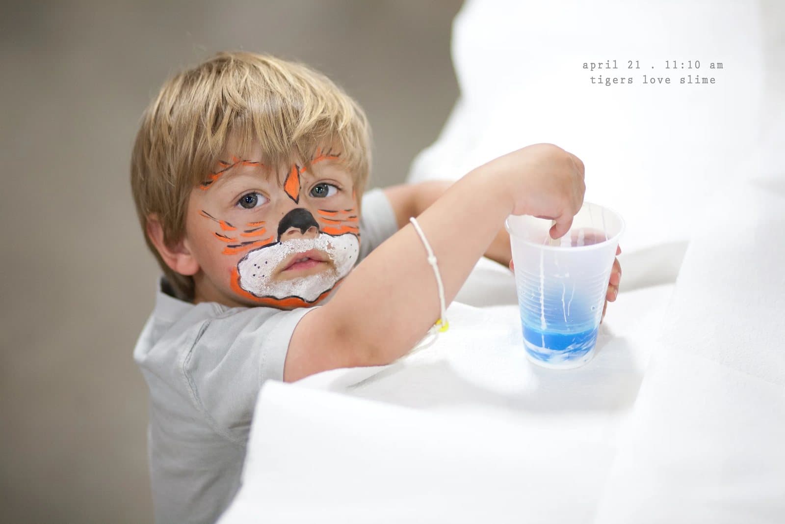 A boy with tiger face paint dips a spoon into a cup of blue slime.
