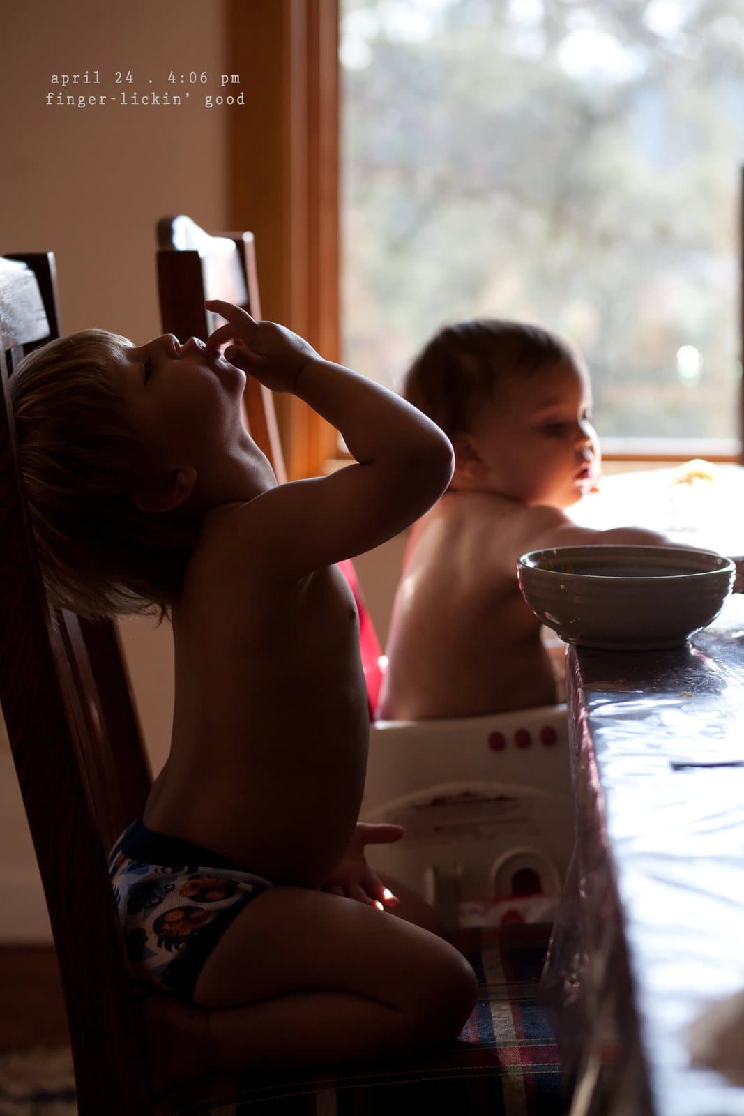A toddler licks his fingers at a sunny table beside a baby, captioned finger-lickin' good.