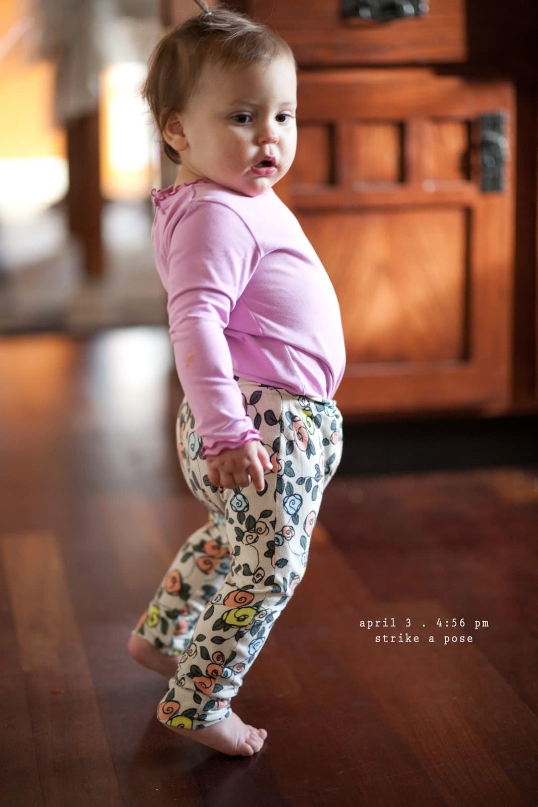 A baby girl in a pink top and patterned pants poses barefoot on a wood floor.
