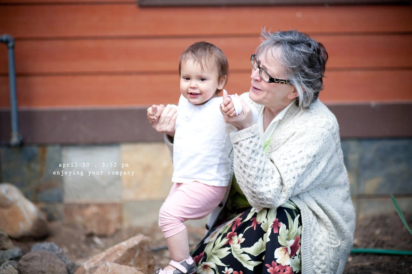 A gray-haired woman holds a smiling baby girl outside a house, captioned enjoying your company.