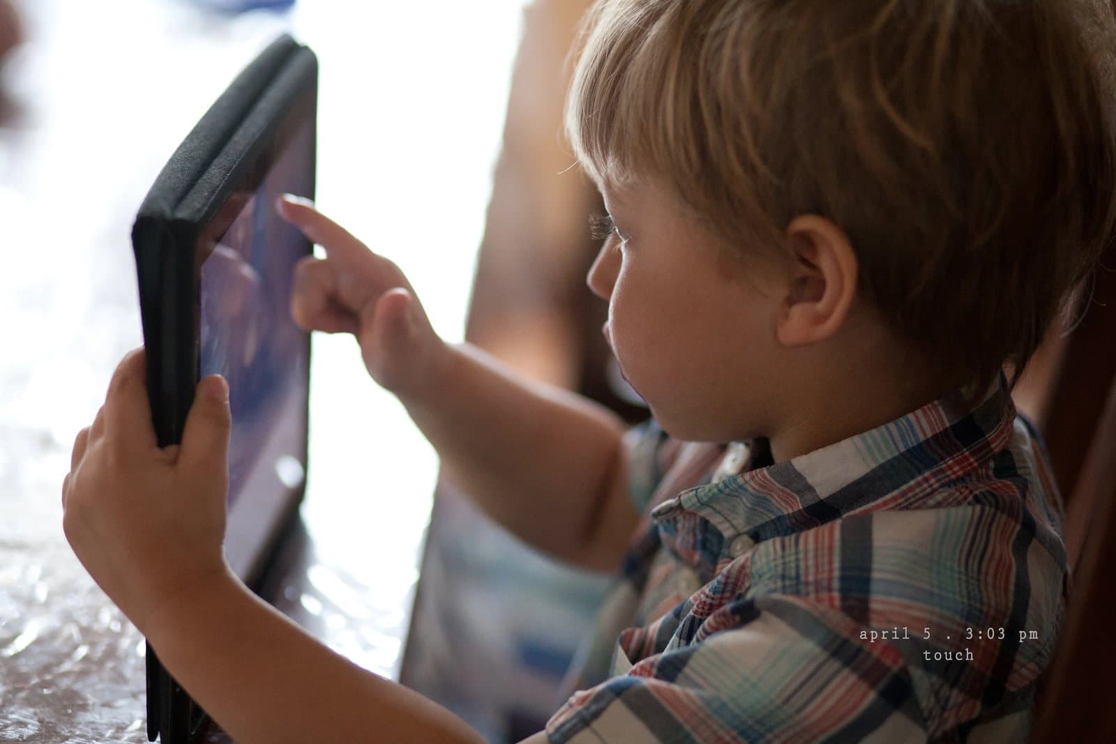 A toddler touches the screen of a tablet in a black case, captioned touch.