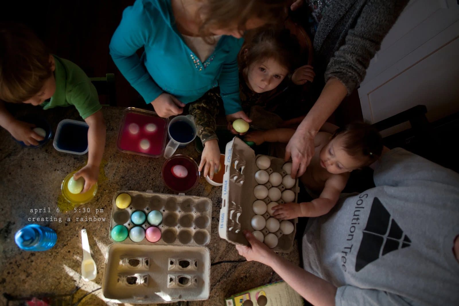 A family dyes Easter eggs in bright colored cups on a table, captioned creating a rainbow.
