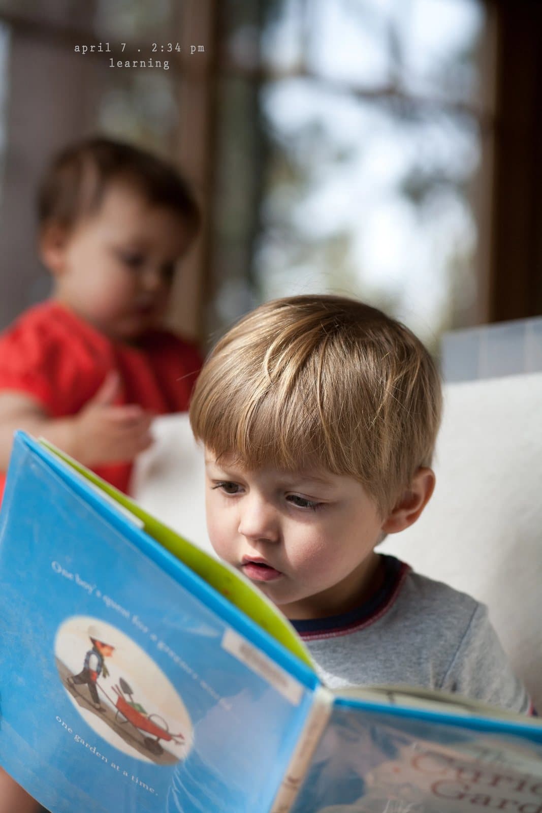 A toddler reads a Curious George picture book while a baby looks on, captioned learning.