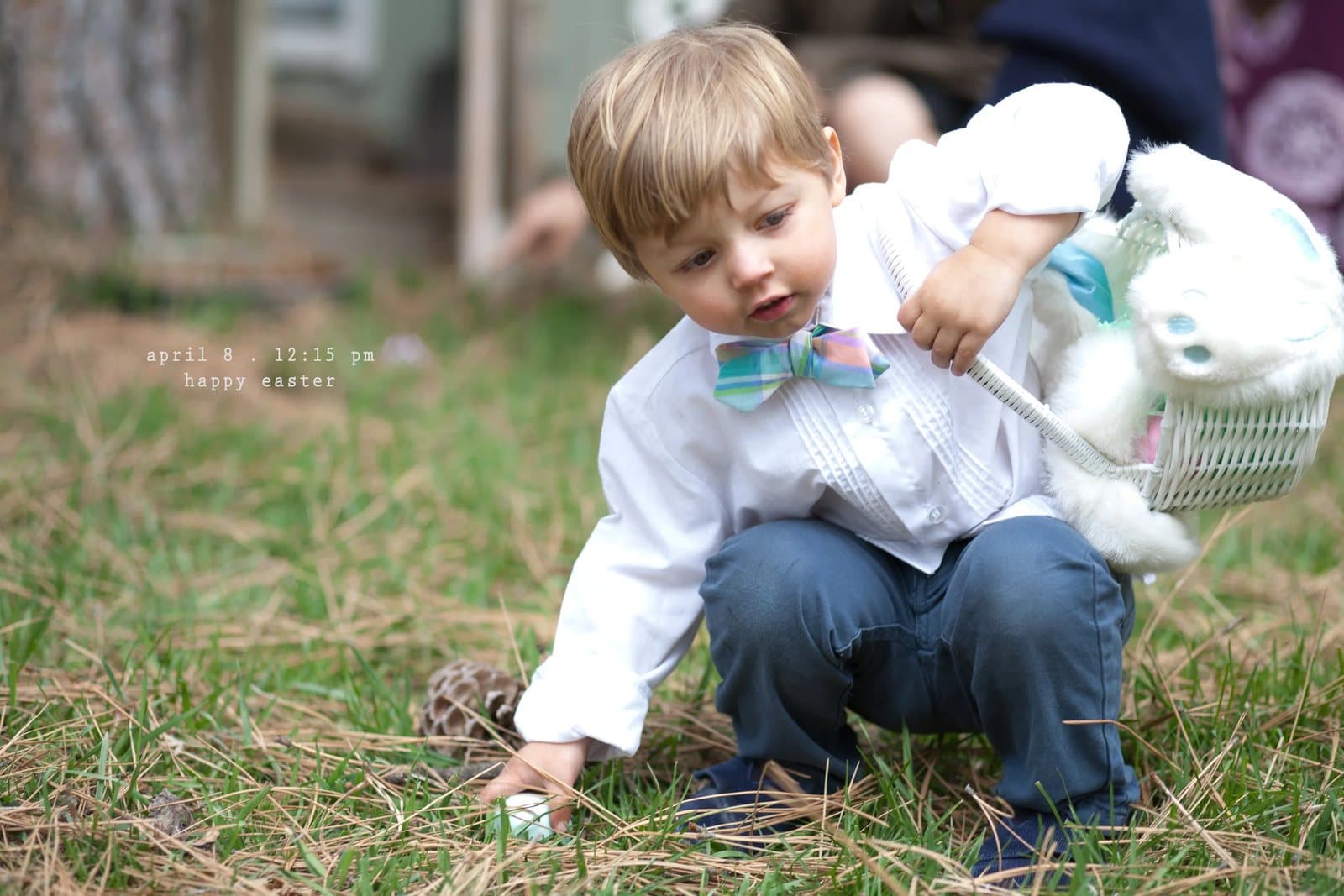 A boy in a bow tie with an Easter basket picks up an egg from the grass.