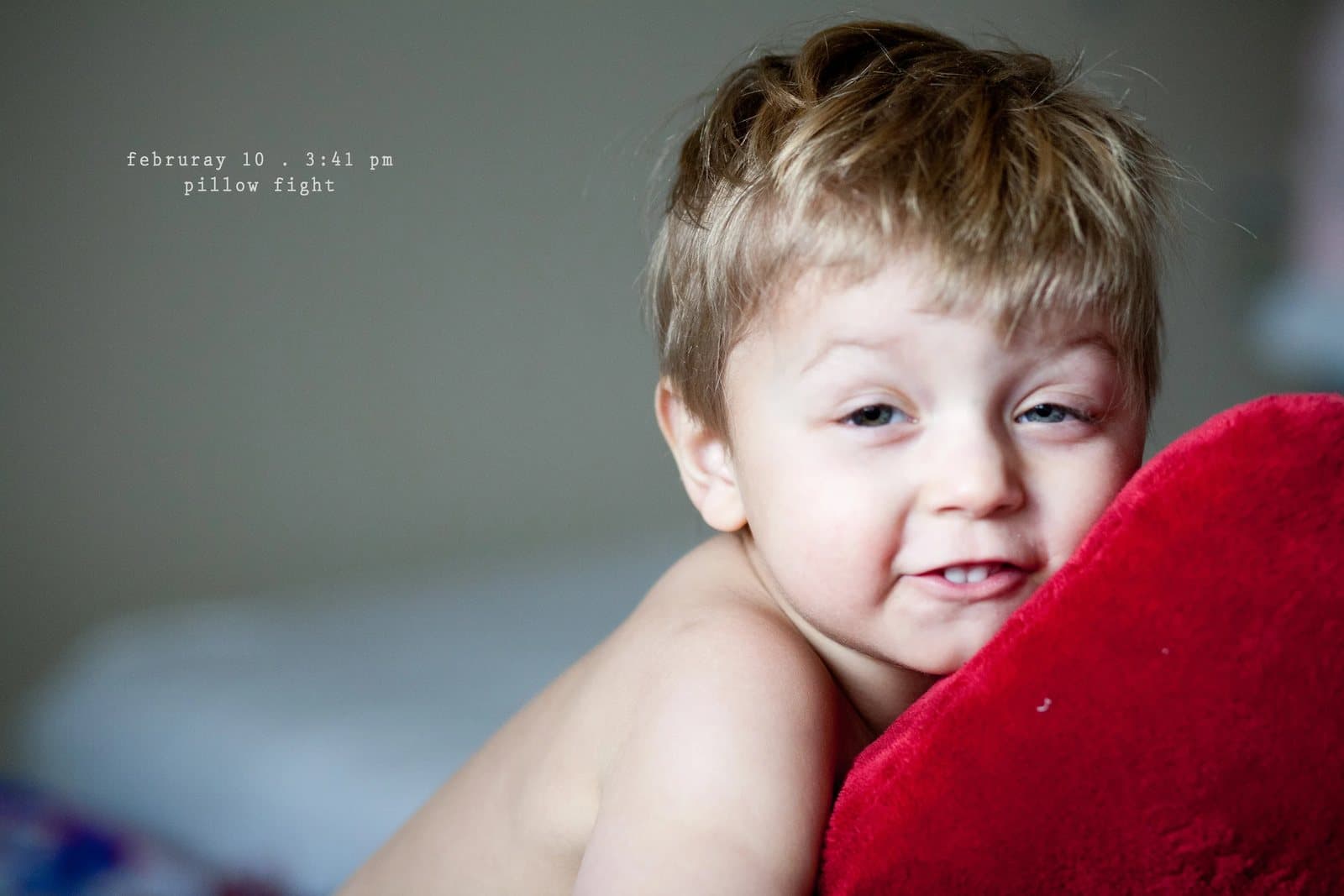 A shirtless toddler grins while hugging a red pillow, captioned pillow fight.
