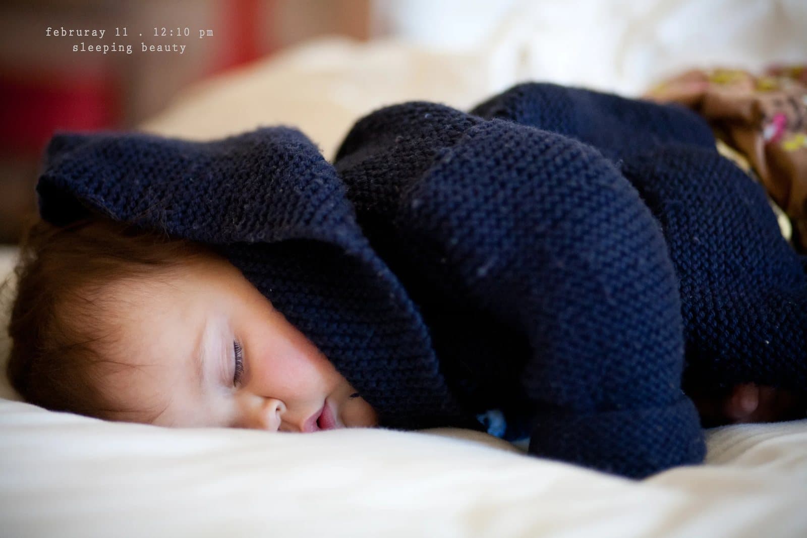 A baby sleeps face down under a knitted navy blanket, captioned sleeping beauty.