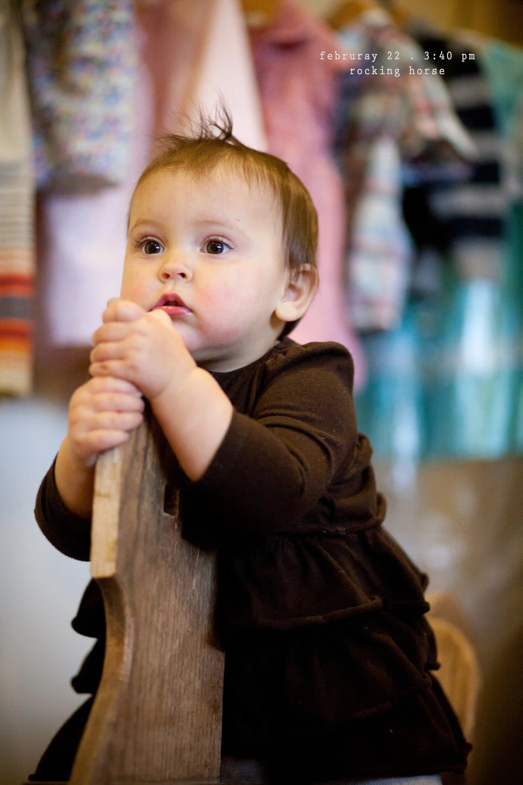 A baby in a brown dress holds on to a wooden rocking horse, captioned rocking horse.