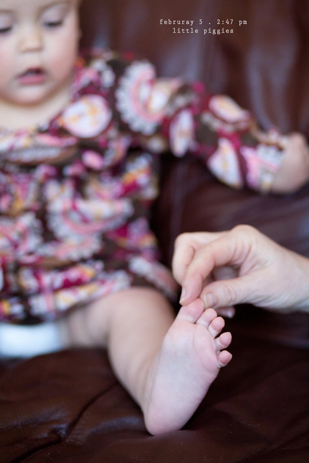 An adult hand holds the bare foot of a baby in a floral outfit, captioned little piggies.