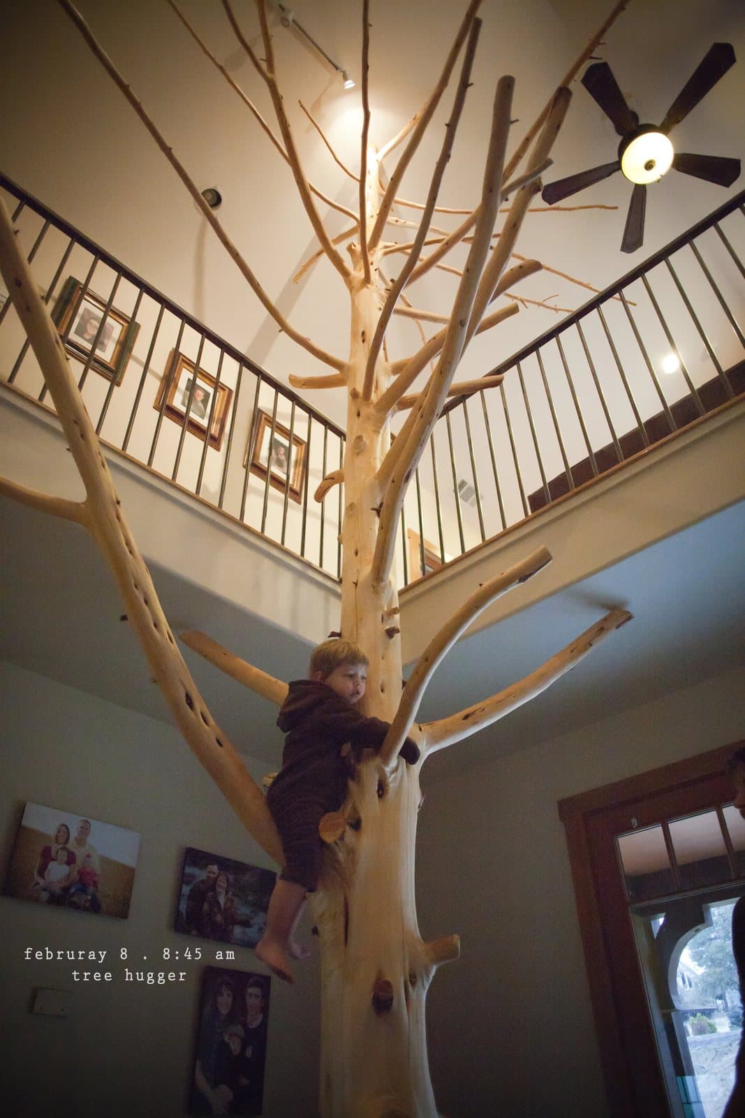 A child climbs a tree-trunk sculpture reaching up to an indoor balcony, captioned tree hugger.