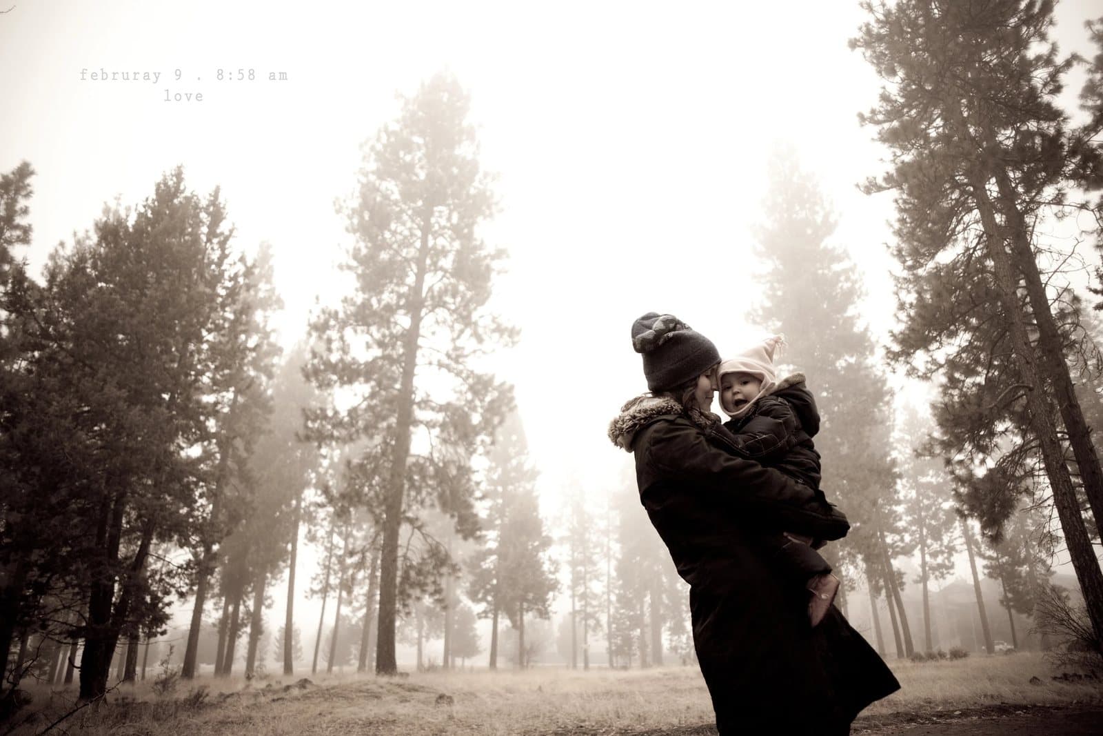A woman holds a child close among tall pines in the fog, captioned love.