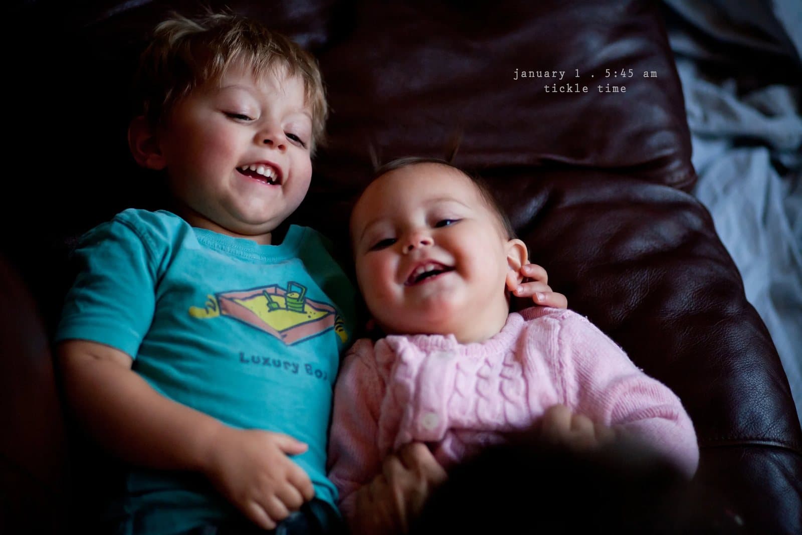 A laughing boy and a baby girl lie on a leather couch, captioned tickle time.