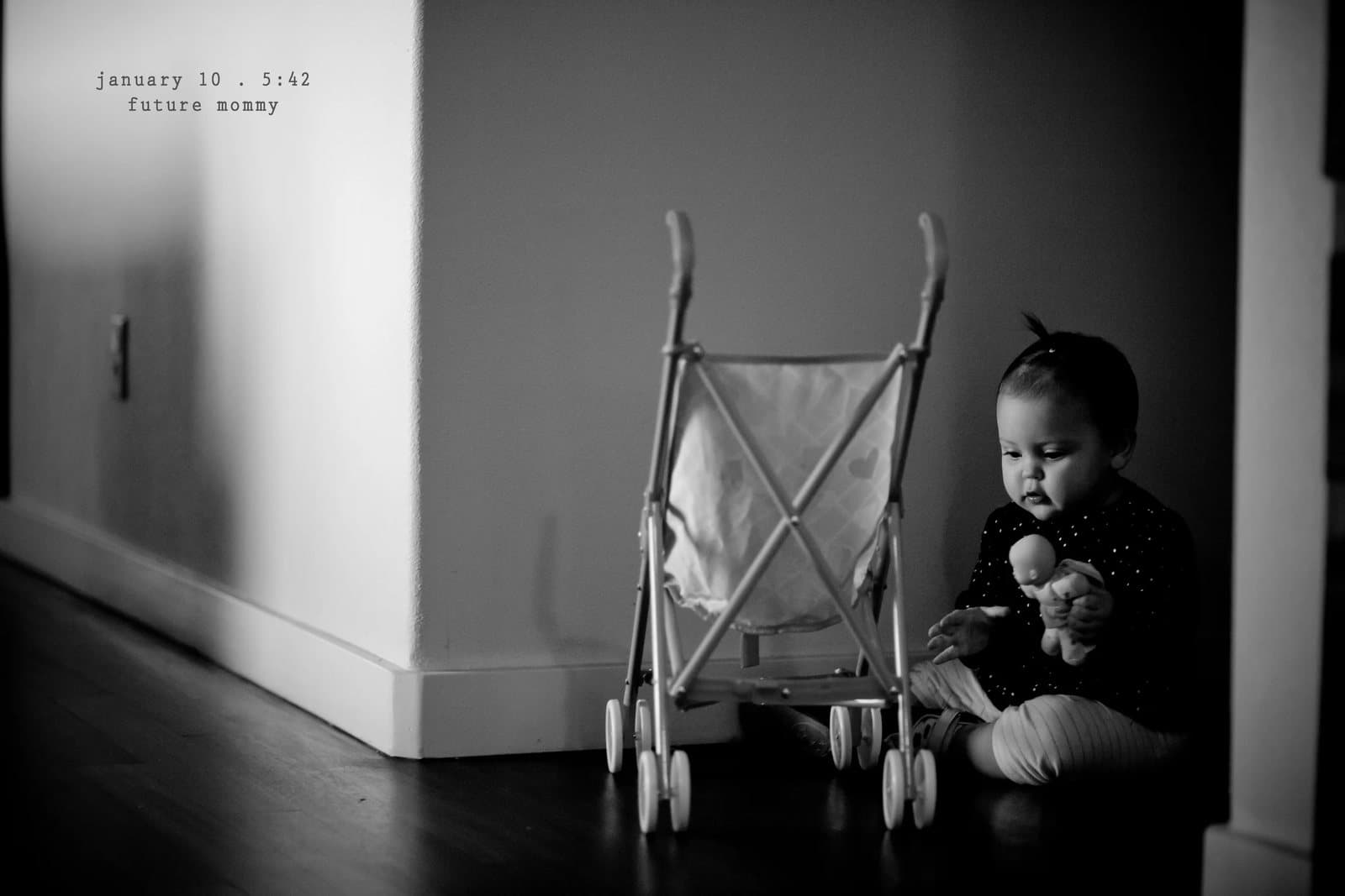 A baby sits beside a toy stroller on a dark wood floor, captioned future mommy.
