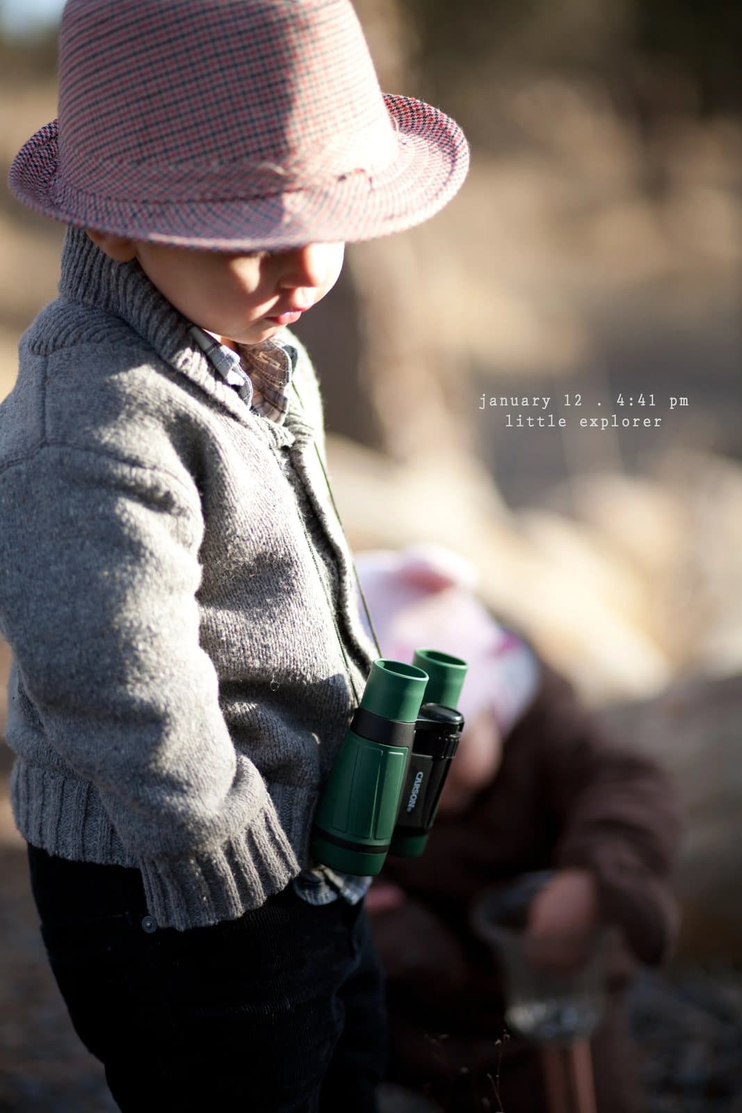 A toddler in a hat and sweater carries green binoculars, captioned little explorer.
