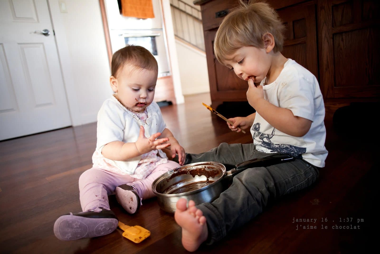 A baby and a toddler lick chocolate from a mixing bowl on the floor, captioned j'aime le chocolat.