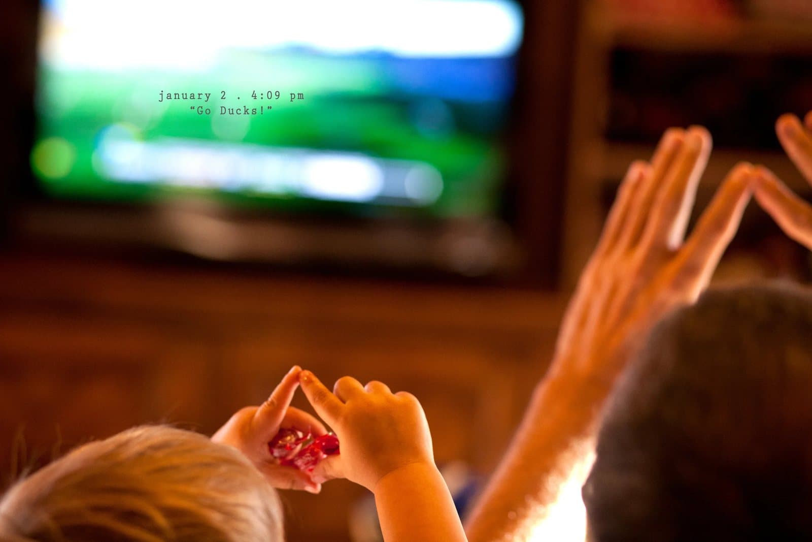 Two children's hands make heart shapes toward a TV showing football, captioned Go Ducks!