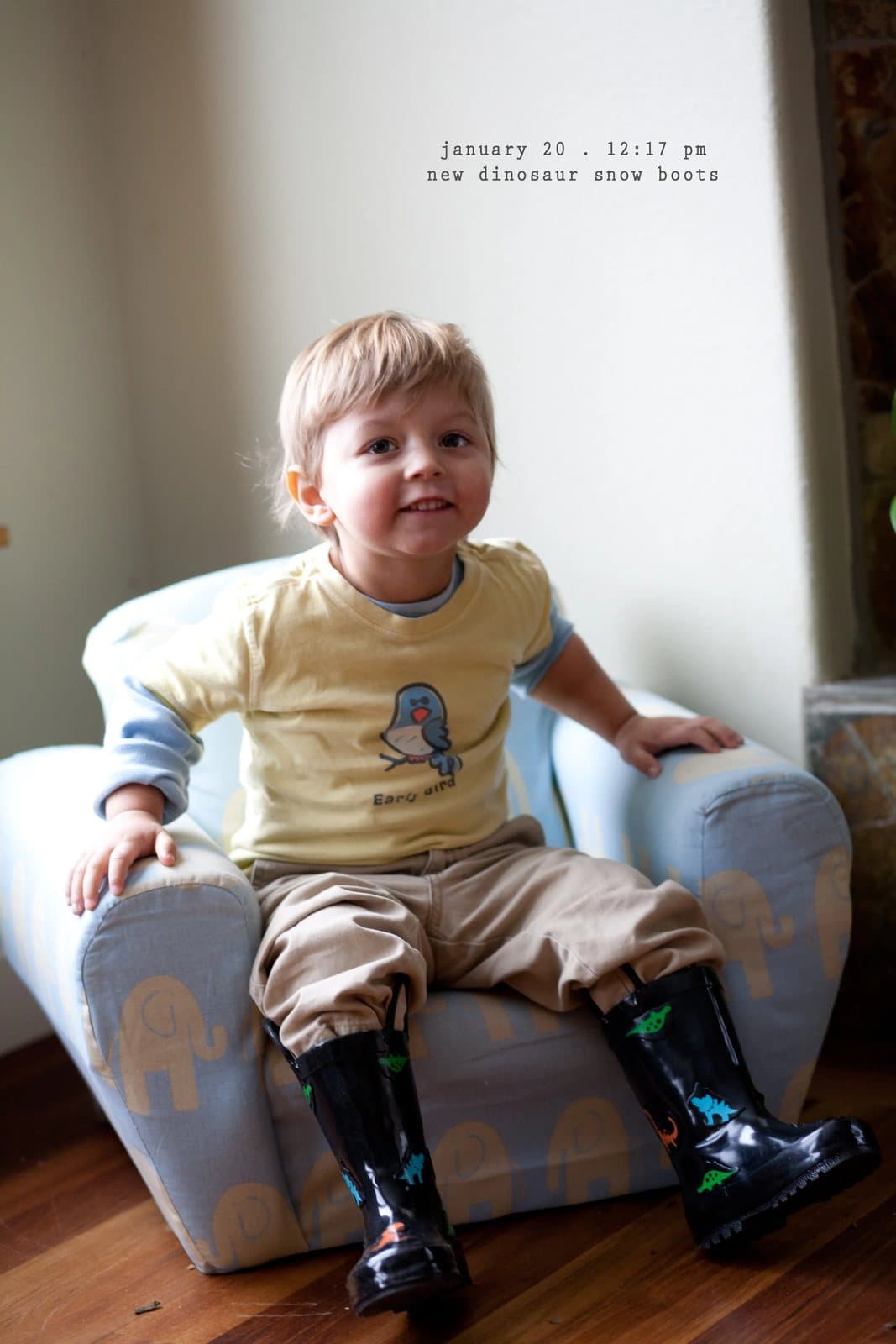 A toddler in dinosaur rain boots sits in a small armchair, captioned new dinosaur snow boots.