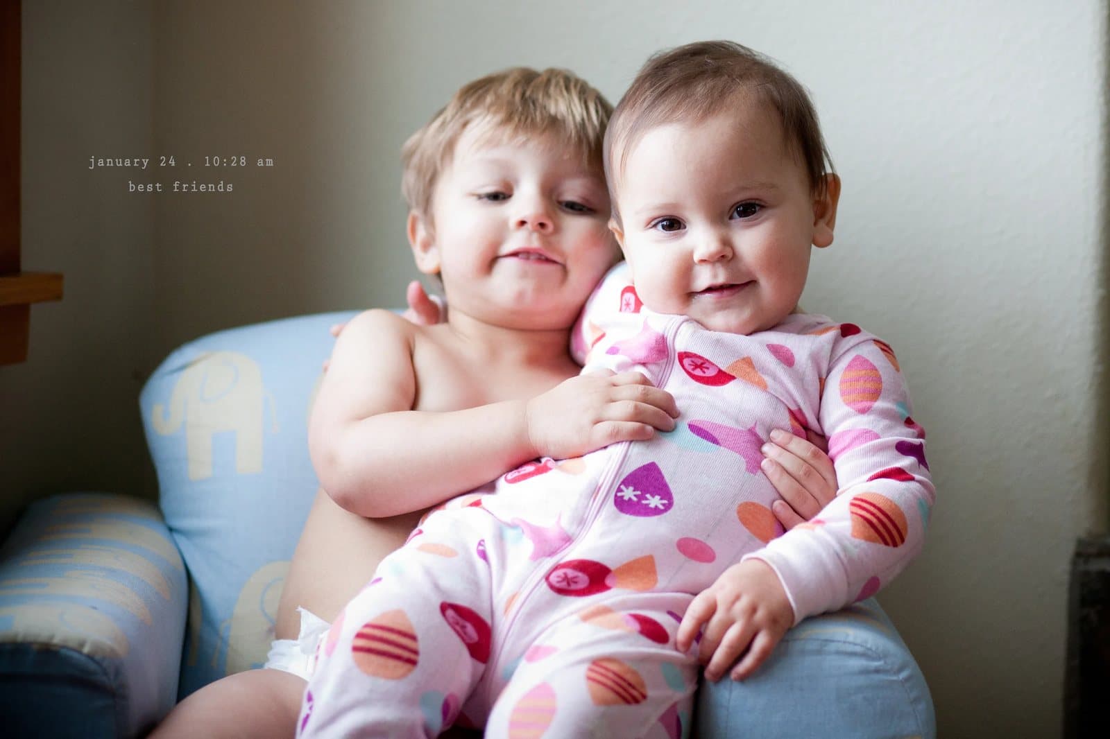 A shirtless toddler hugs a smiling baby in pajamas on an armchair, captioned best friends.