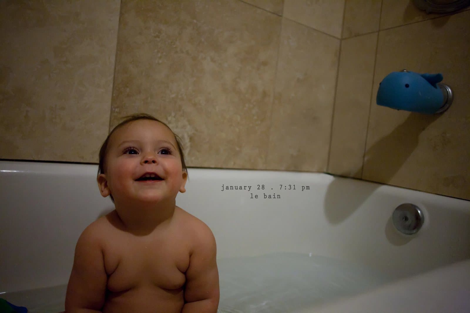 A smiling baby sits in a bathtub below a whale spout cover, captioned le bain.