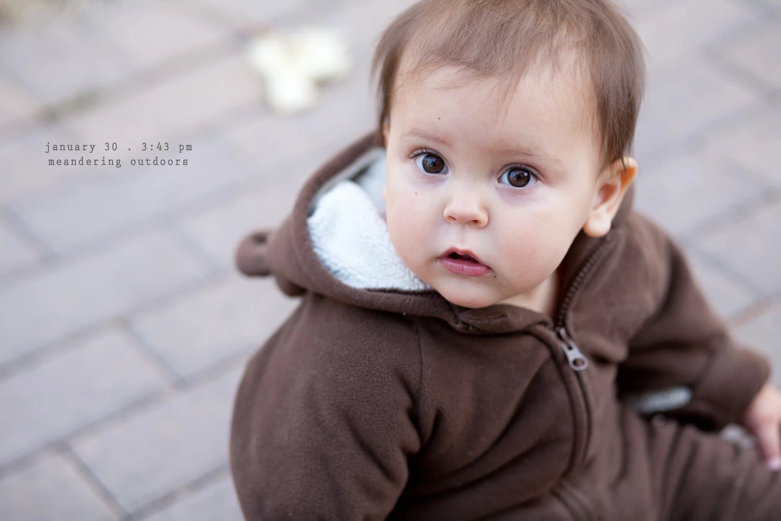 A baby in a brown fleece hoodie sits on pavers looking up, captioned meandering outdoors.