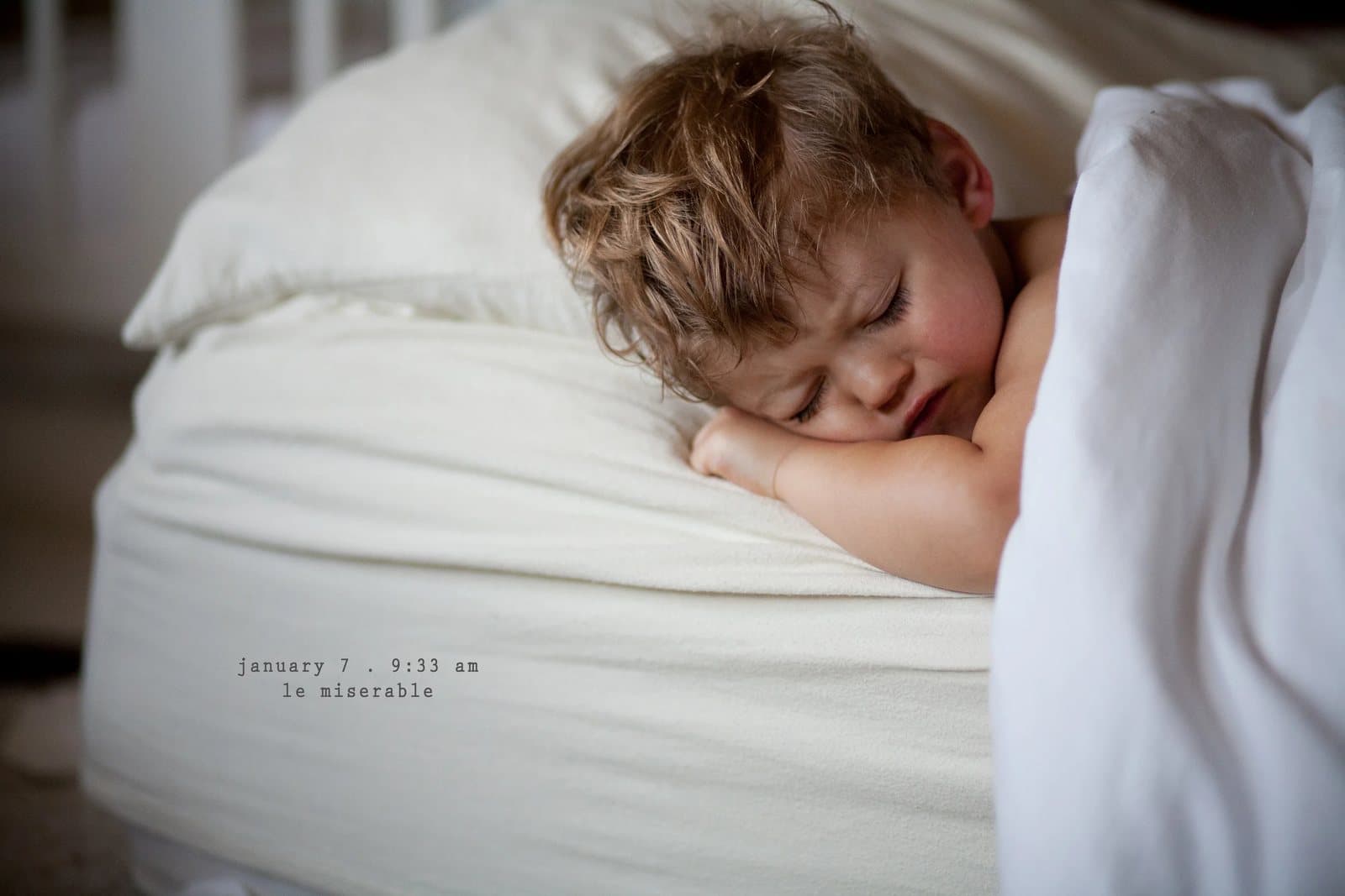 A tearful toddler lies on a white pillow in bed, captioned le miserable.