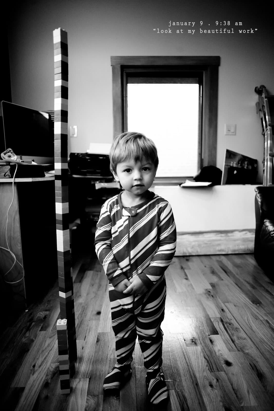 A toddler in striped pajamas stands beside a tall block tower, captioned look at my beautiful work.