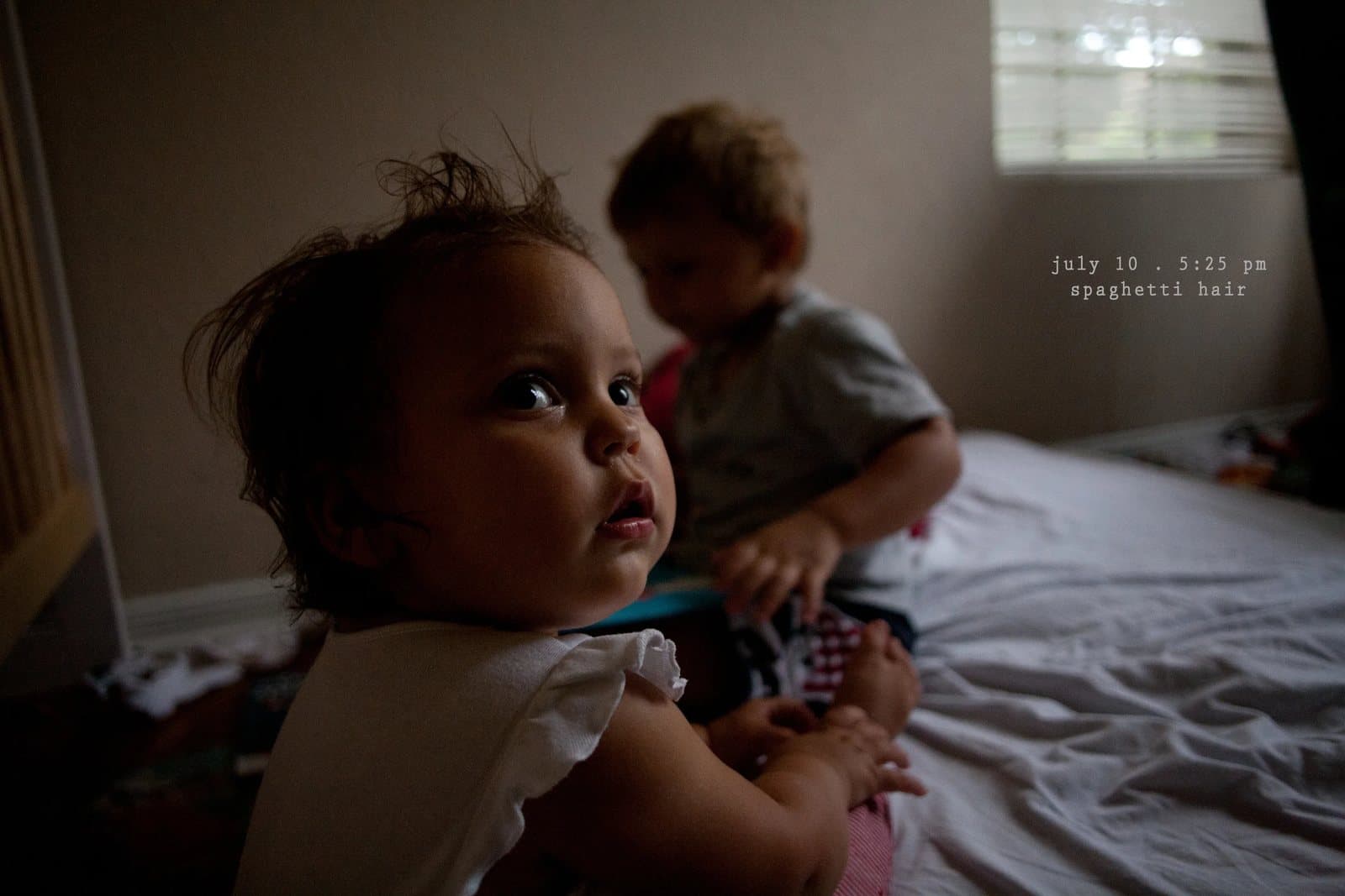 A baby looks back from a bed as a toddler plays behind her, captioned spaghetti hair.