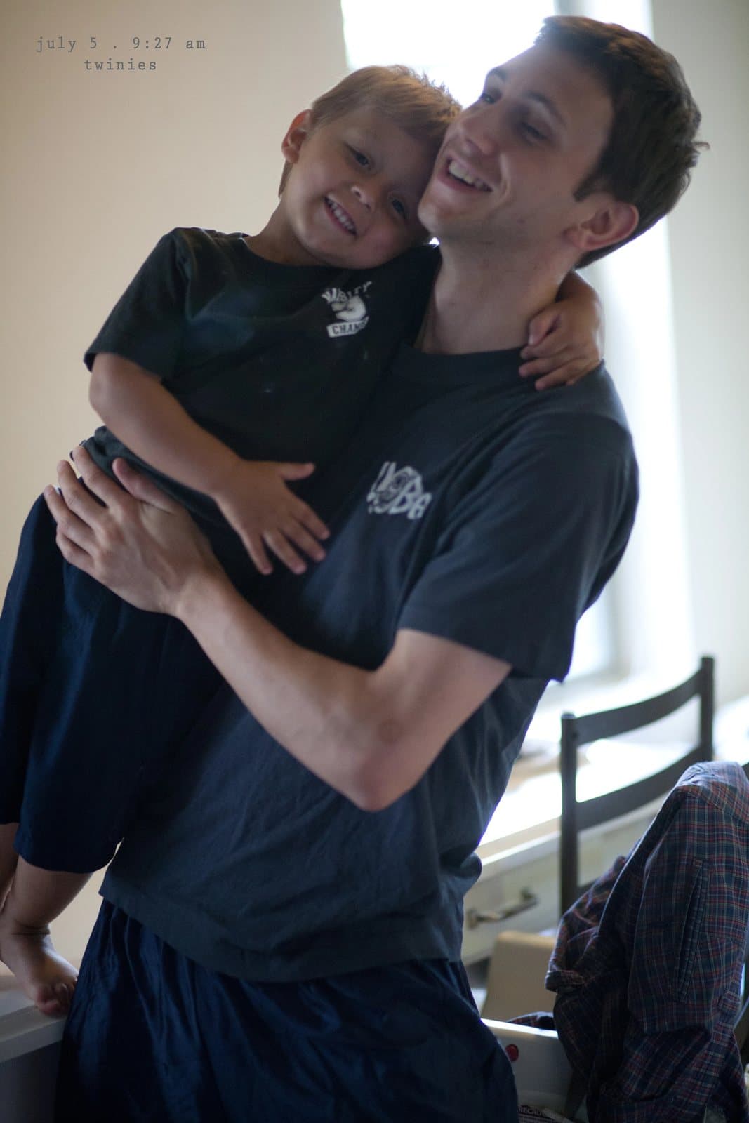 A smiling young man carries a laughing boy in matching navy shirts.
