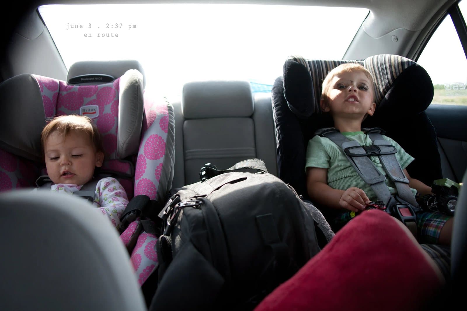A baby and a toddler ride in car seats in a back seat, captioned en route.