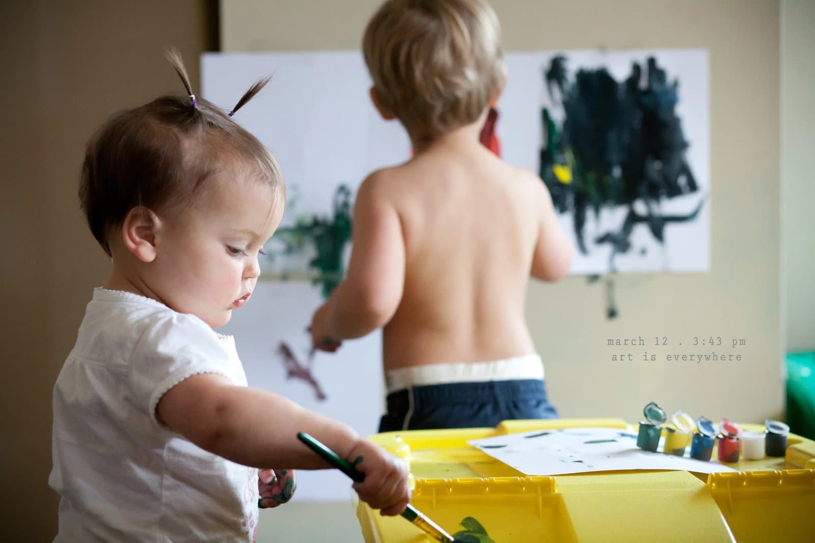 A baby and a shirtless toddler paint at an easel, captioned art is everywhere.