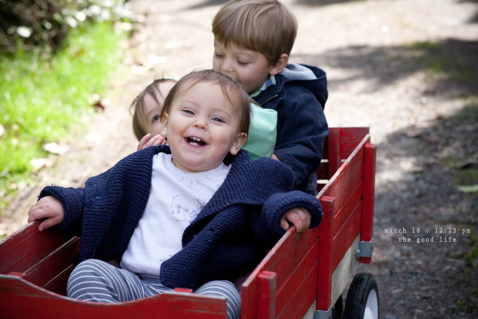 Three laughing children ride in a red wagon on a trail, captioned the good life.