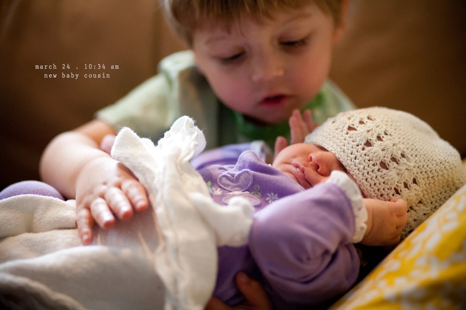 A toddler cradles a sleeping newborn in a knit cap, captioned new baby cousin.