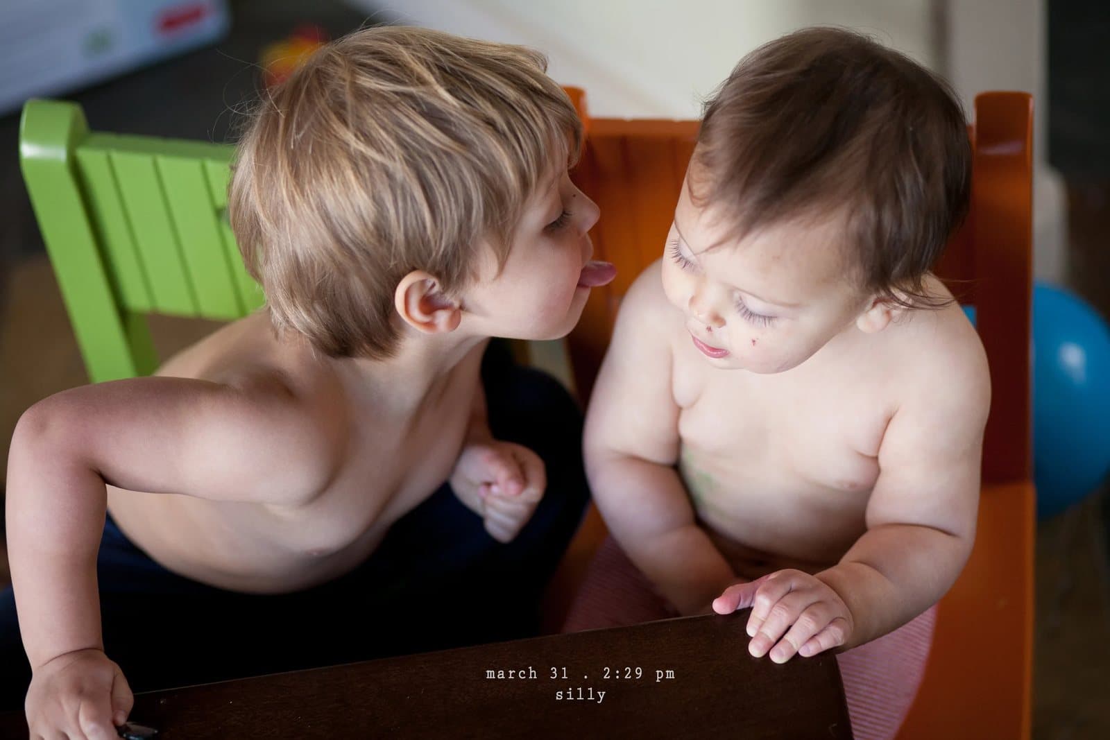A shirtless boy sticks out his tongue at a giggling baby, captioned silly.