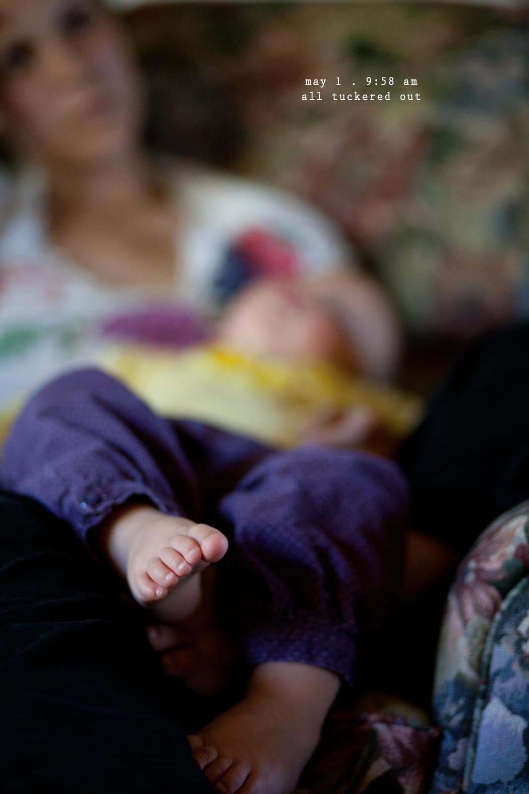 A sleeping baby's bare feet rest on a woman's lap, captioned all tuckered out.