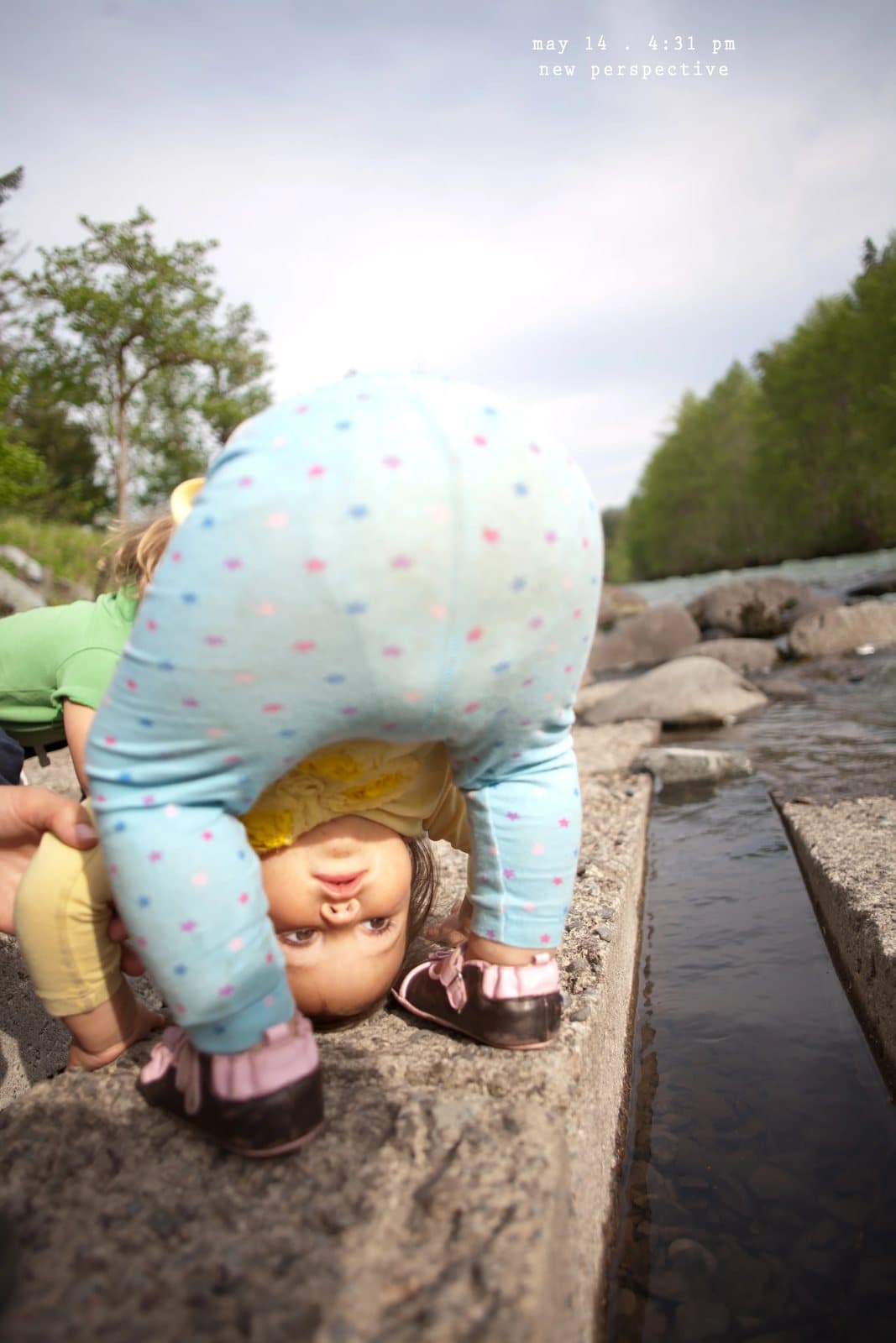 A child bends over to look upside down between her legs by a creek, captioned new perspective.