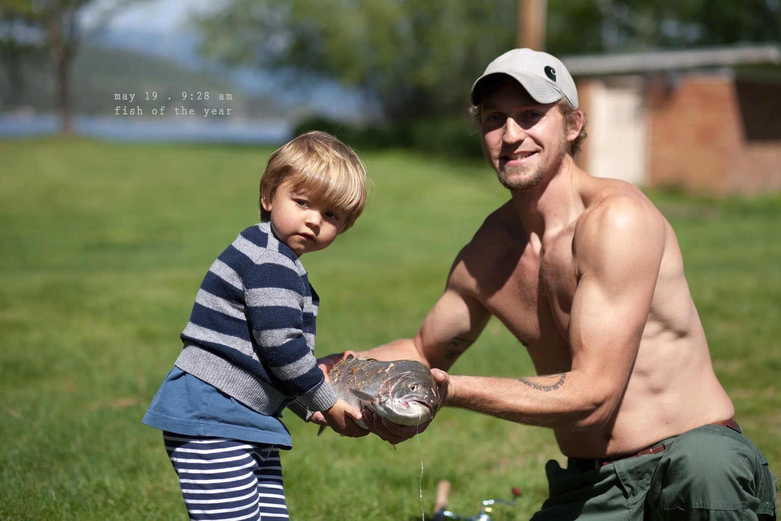 A shirtless man and a toddler hold up a large trout on a lawn, captioned fish of the year.