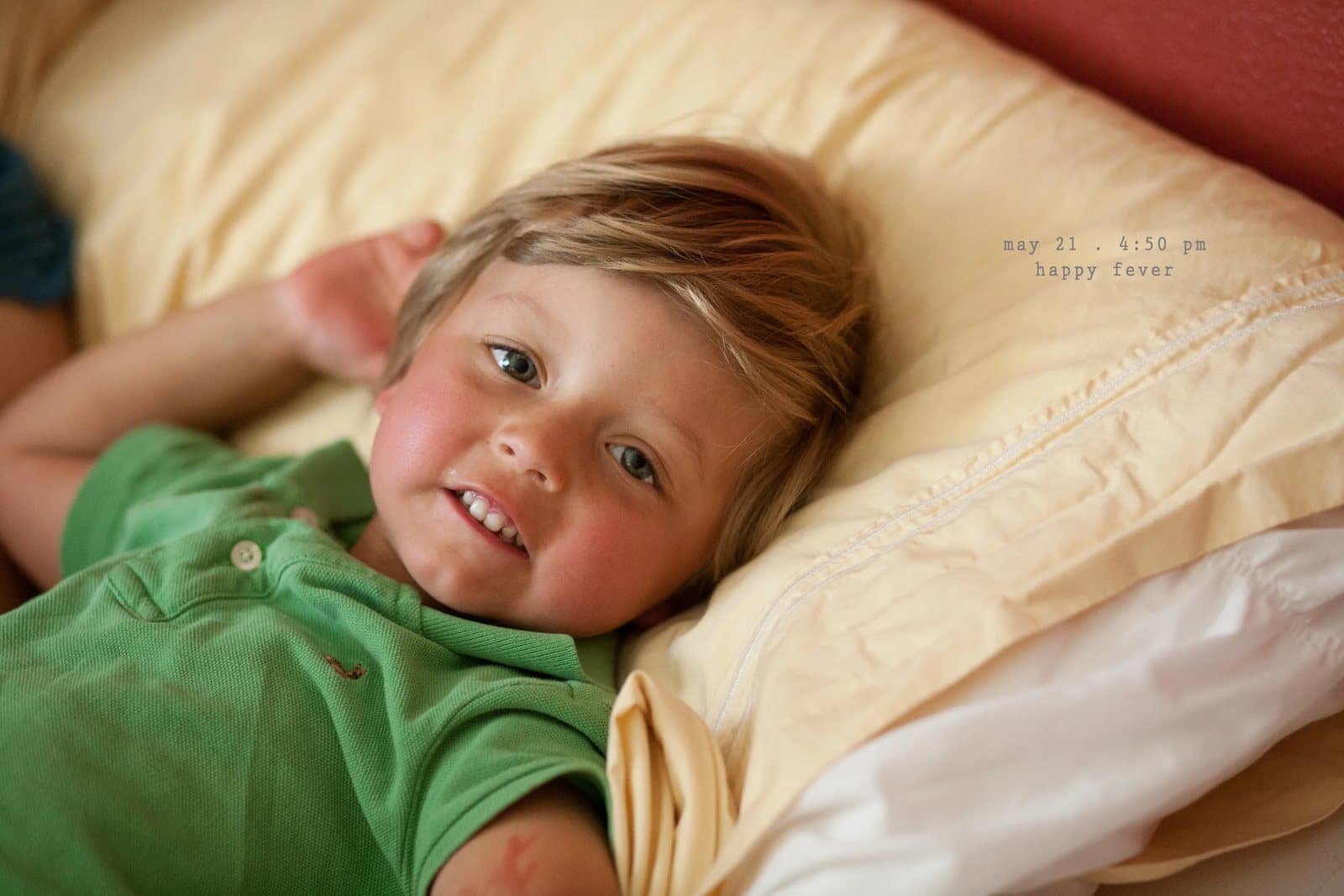 A flushed toddler lies on a pillow smiling, captioned happy fever.