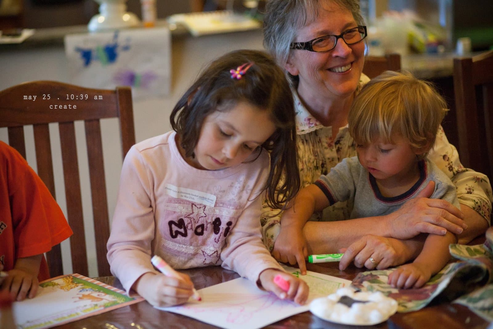 A grandmother draws with a girl and a toddler at a table, captioned create.