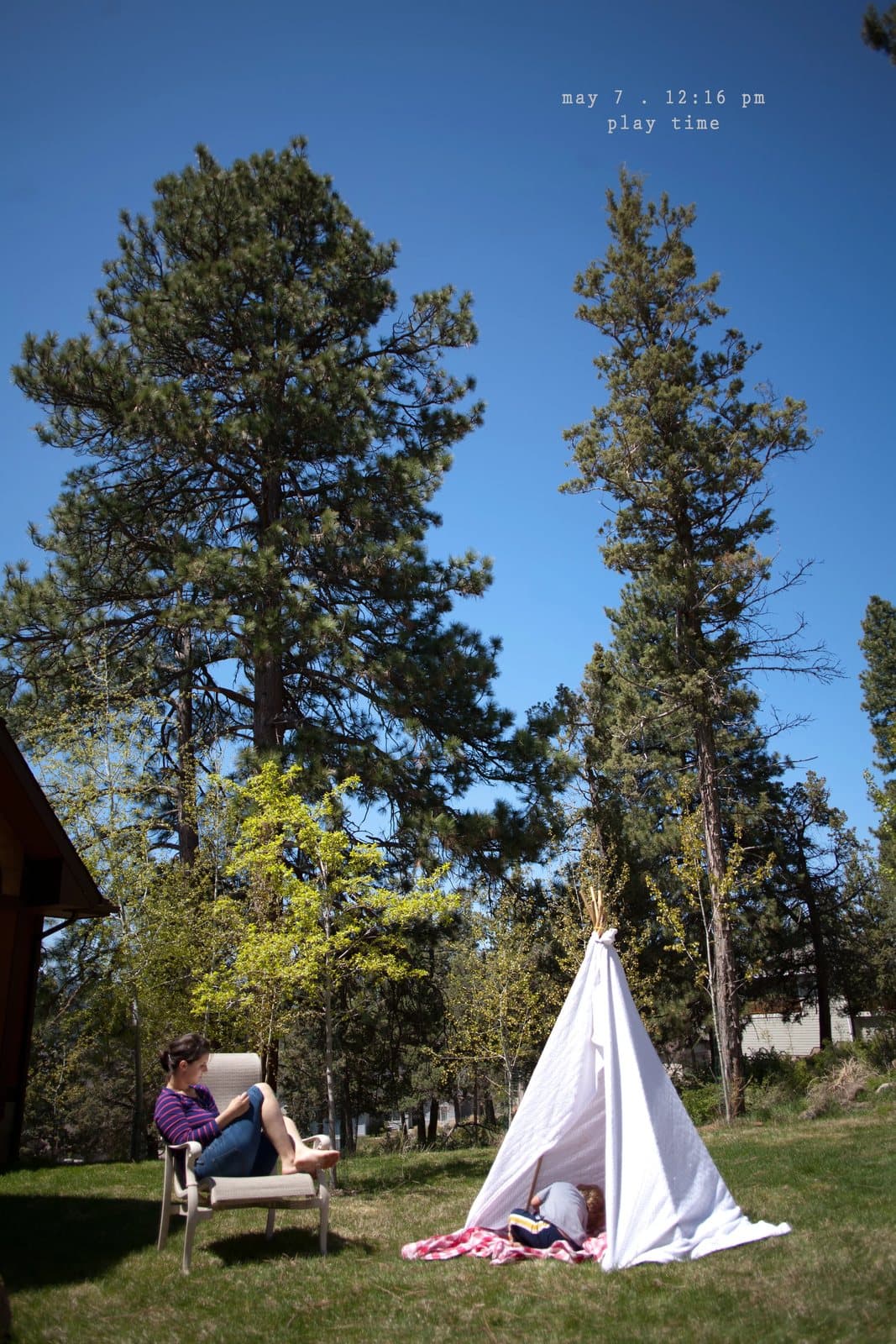 A woman reads in a chair beside a white teepee under tall pines, captioned play time.