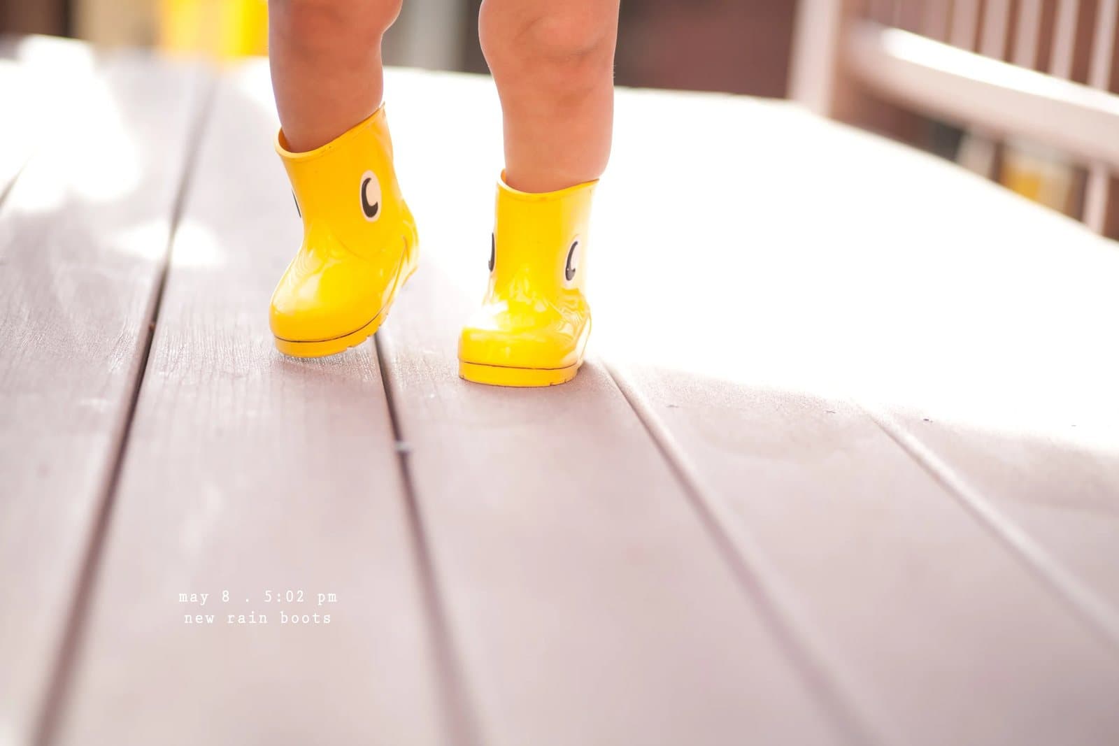 A toddler's legs in yellow rain boots on a wooden deck, captioned new rain boots.