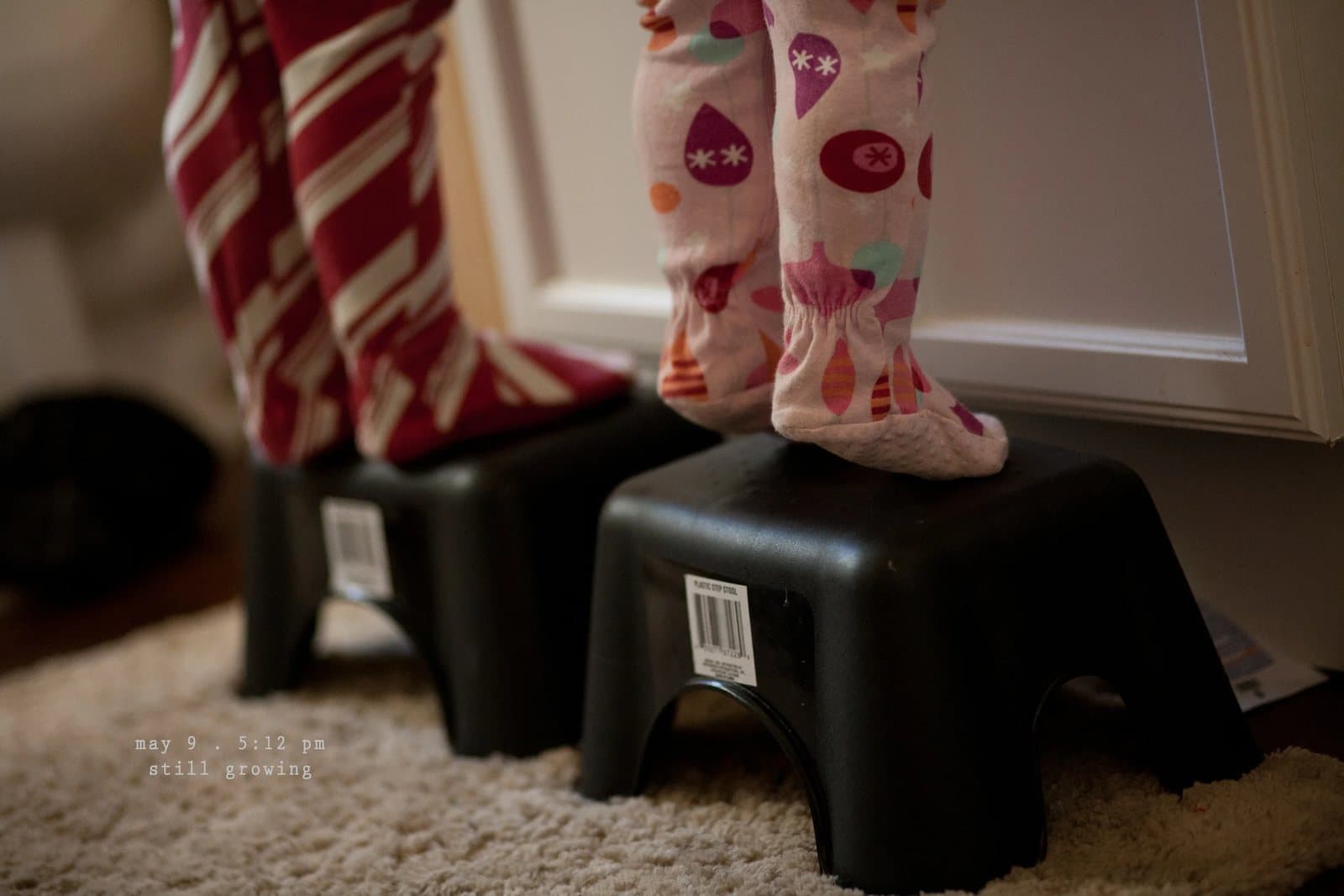Two children in pajamas stand on black step stools, captioned still growing.