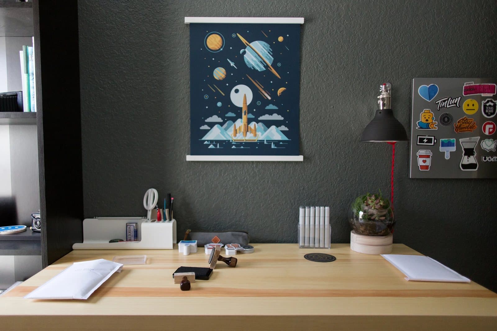 Kyle's airy workspace with a space poster, black lamp, terrarium, and stickered laptop on the wall.