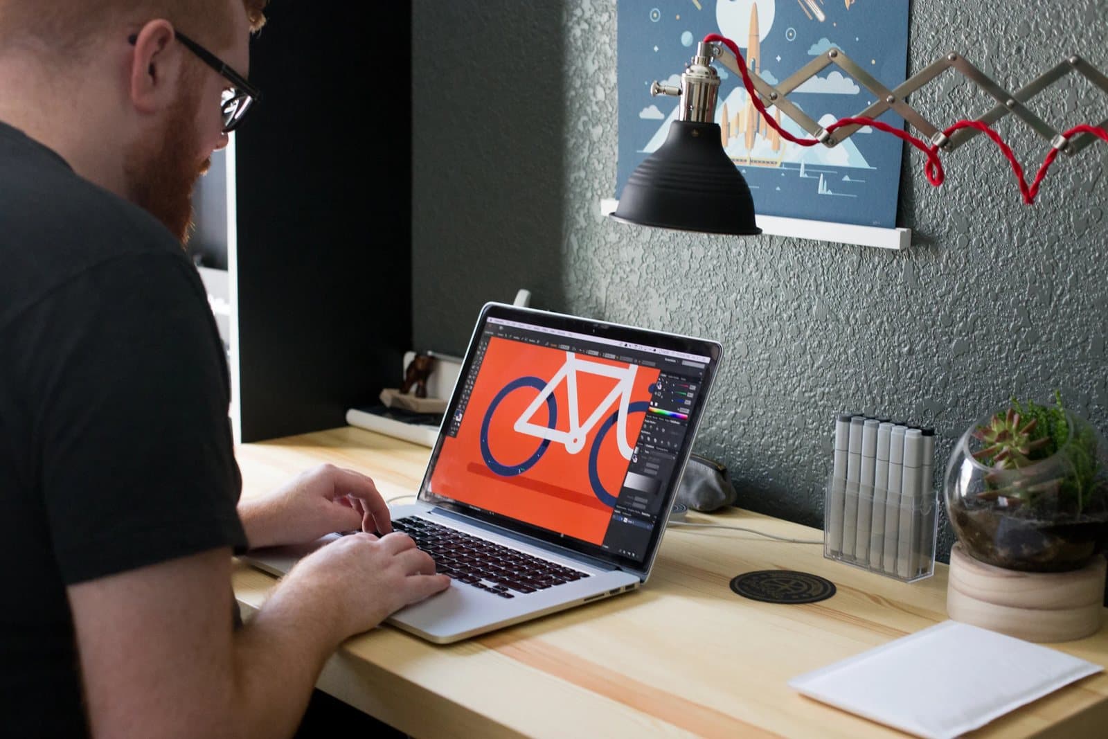 Over Kyle's shoulder, a laptop shows a bicycle icon being designed at his pine desk.