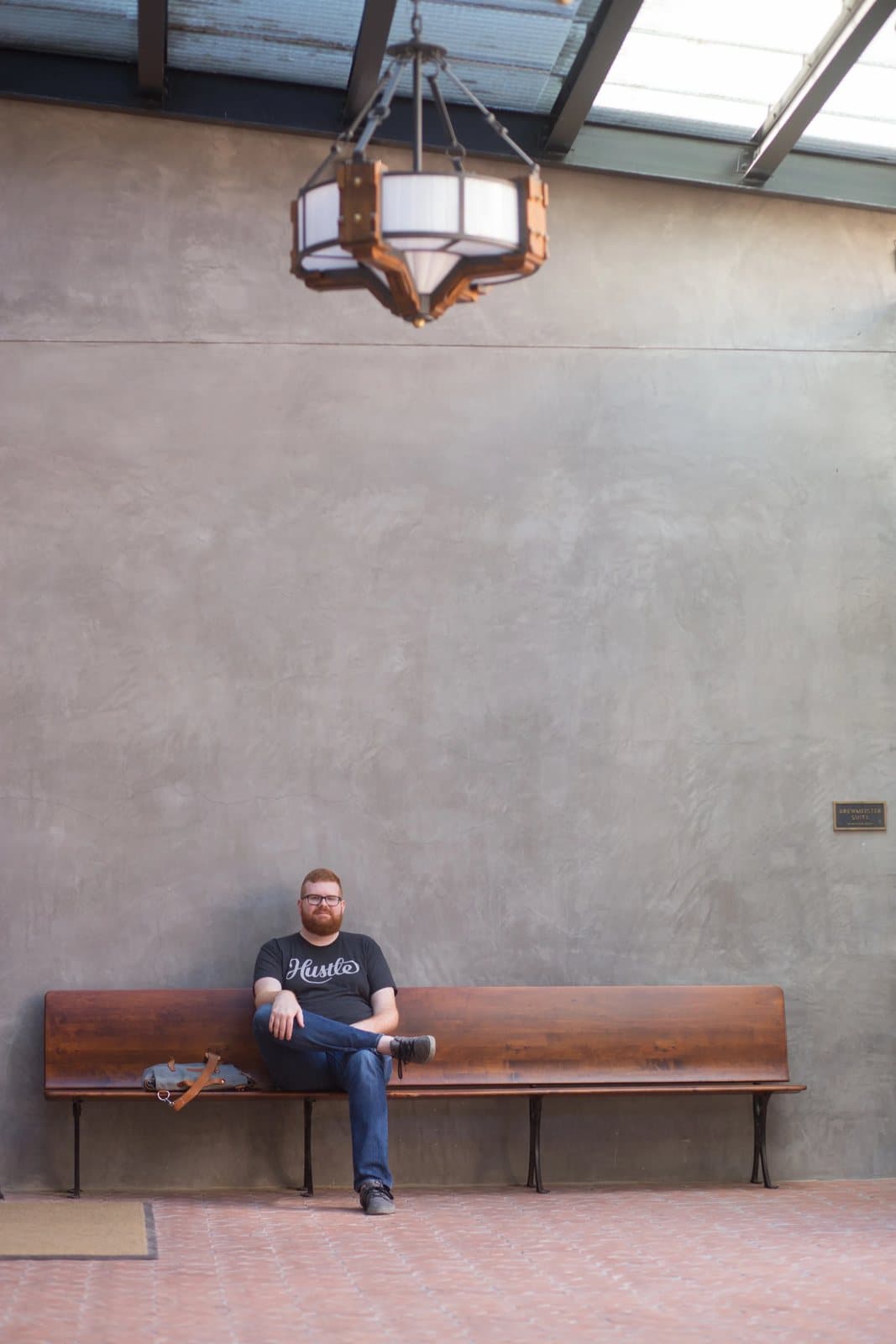 Kyle sits on a long wooden bench beneath a hanging lantern in a skylit plaster hall.
