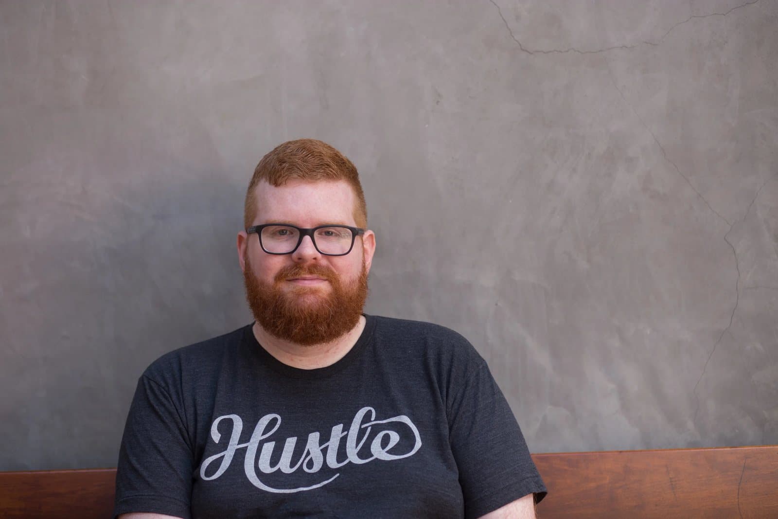 Close portrait of Kyle in glasses and a Hustle T-shirt against a gray plaster wall.