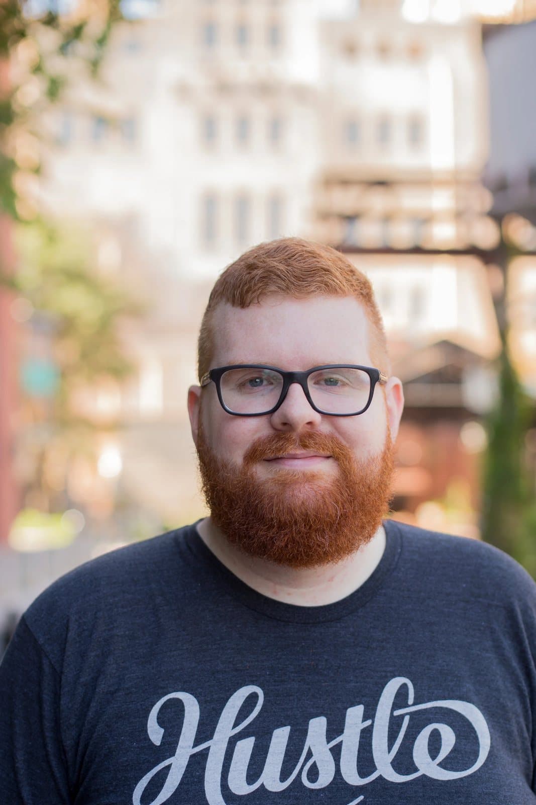 Kyle smiles softly in a Hustle T-shirt with blurred buildings of the Pearl behind him.