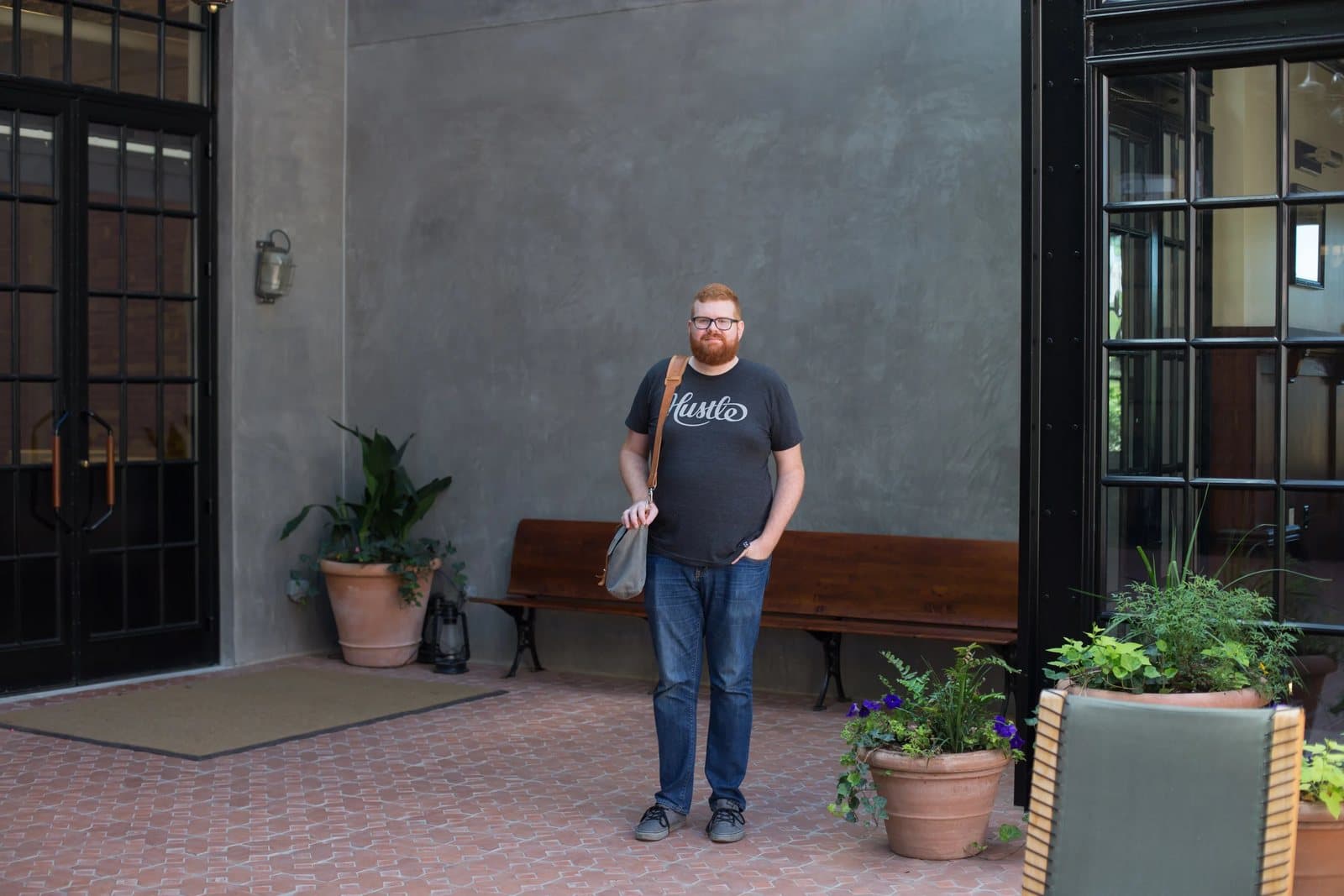 Kyle stands with his bag in a brick courtyard framed by black doors, plants, and a bench.