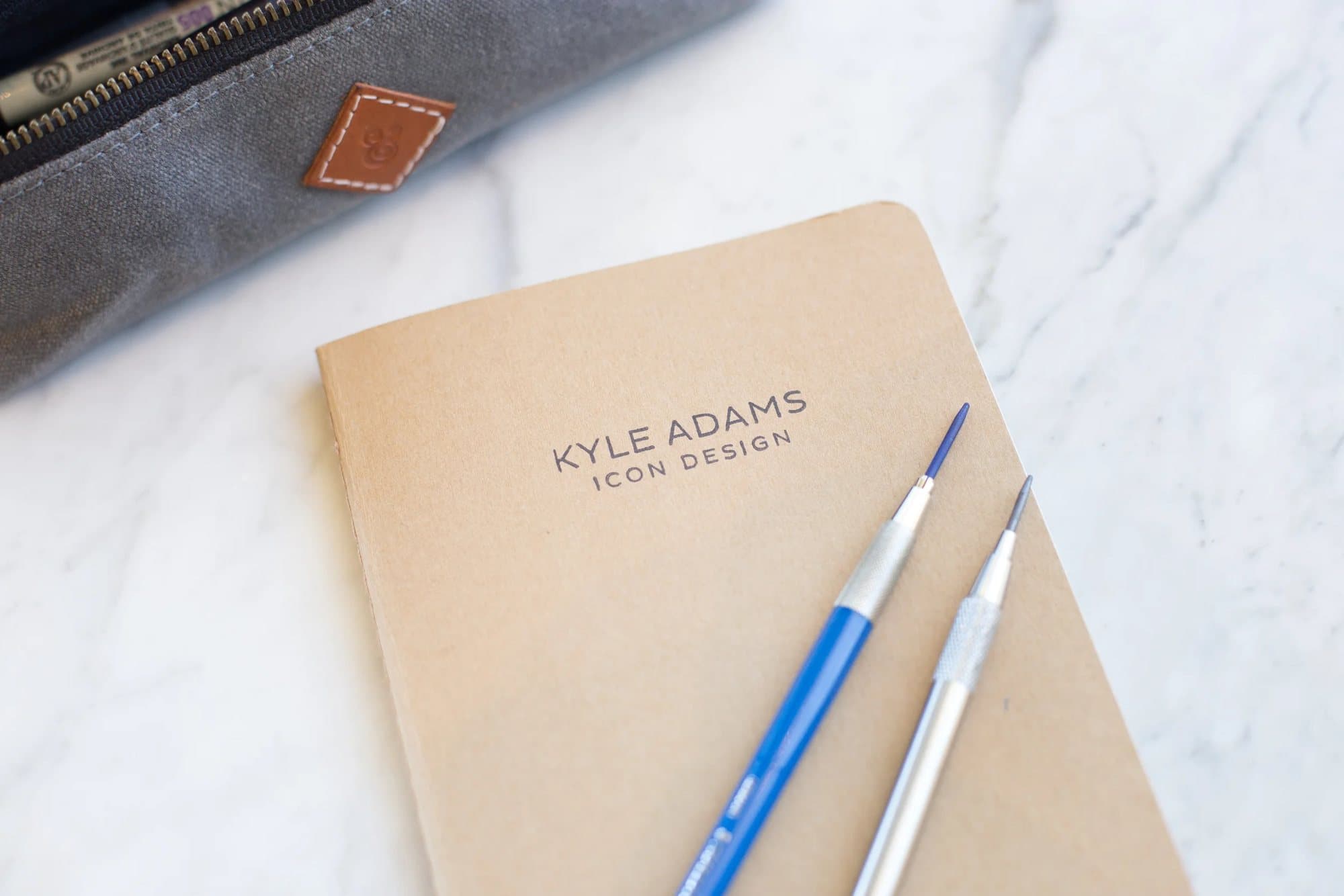 A kraft notebook printed Kyle Adams Icon Design lies on marble with two mechanical pencils.