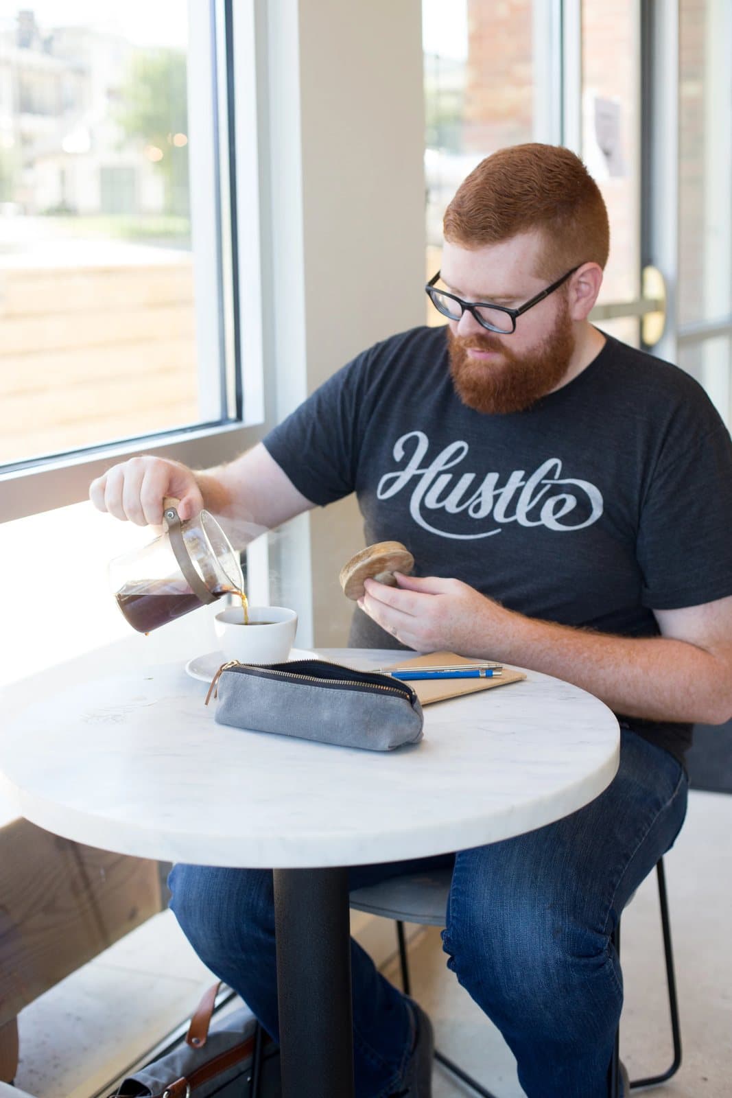 Kyle pours coffee from a carafe at a sunny cafe window table, his pencil case nearby.